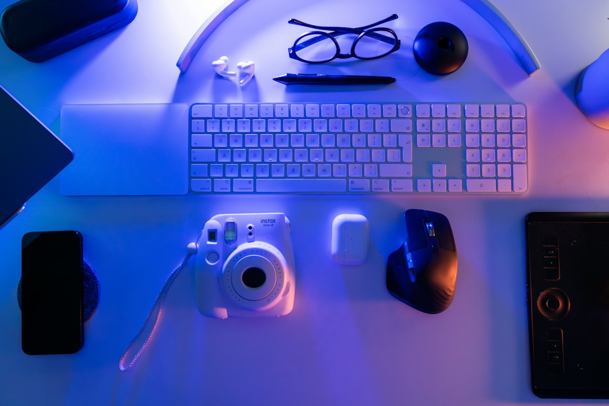 A desk flat lay of a keyboard, mouse, phone and instant camera lit by blue and violet light
